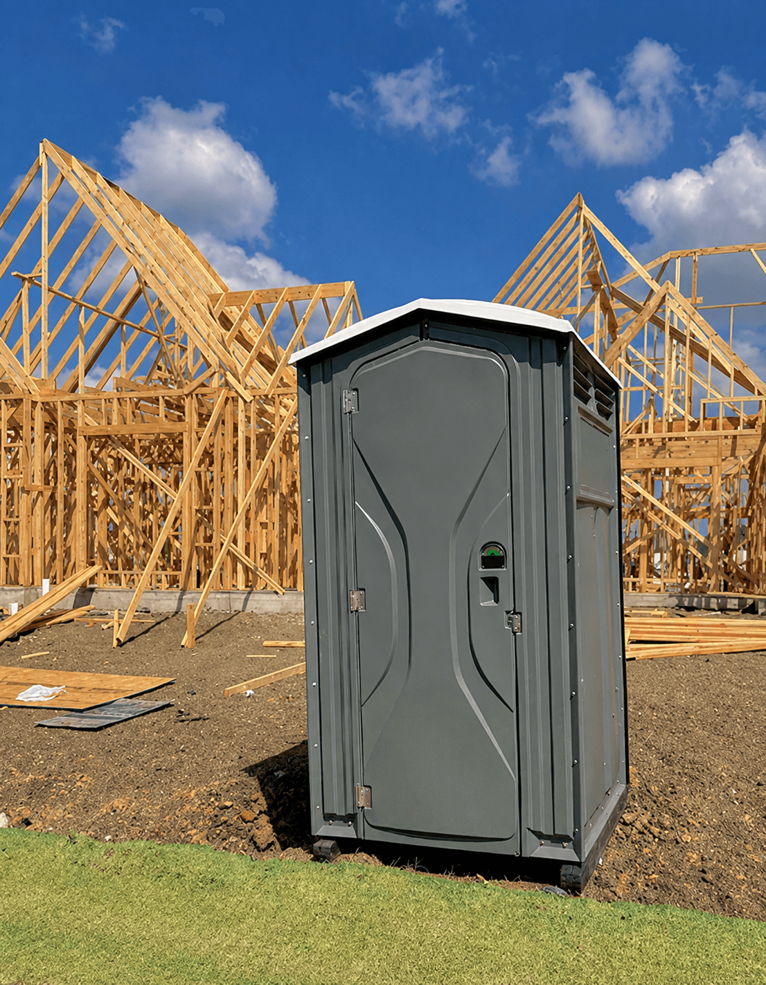Gray portable toilet at a residential construction site with wooden house frames under blue sky.