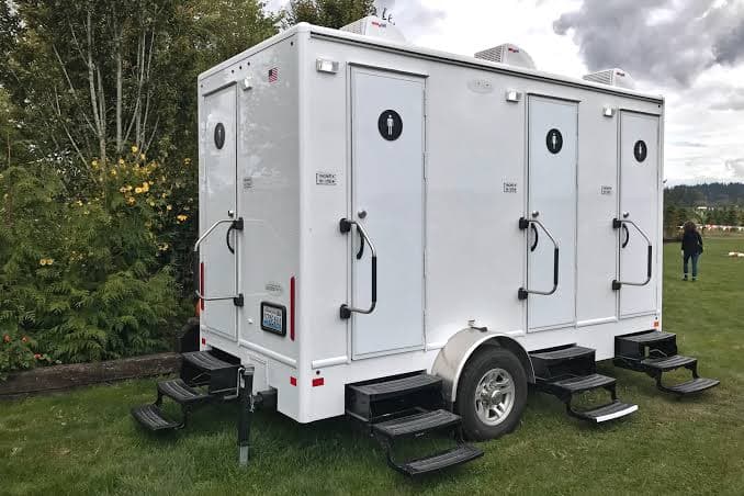 White multi-stall portable restroom trailer with black entry steps parked outdoors on a grassy lawn.